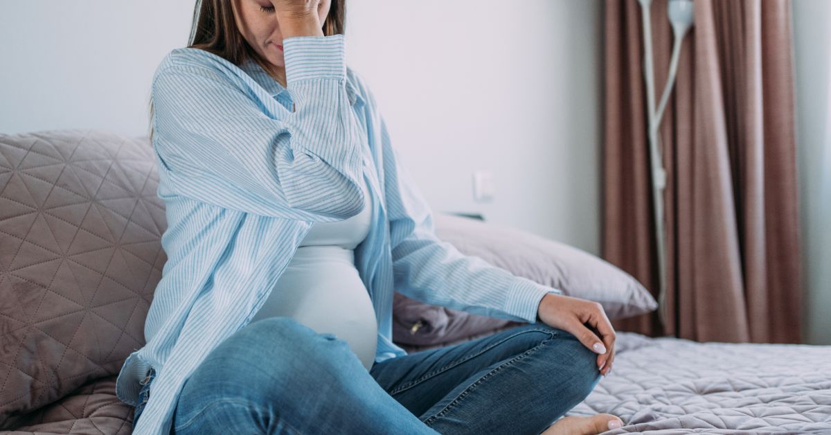 Fotografija trudnice u plavoj odeći koja sedi na krevetu i drži se za glavu